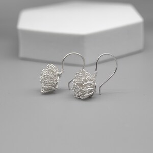 May include: A pair of silver wire earrings with a woven, textured design. The earrings have a hook closure and a cluster of woven wire. The background is a light gray surface with a white geometric shape.