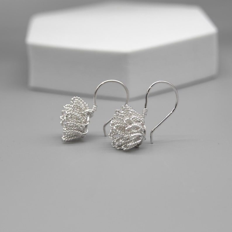 May include: A pair of silver wire earrings with a woven, textured design. The earrings have a hook closure and a cluster of woven wire. The background is a light gray surface with a white geometric shape.