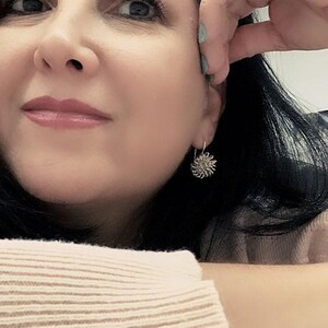 May include: A close-up of a person wearing a silver earring. The earring is a small, round design with a textured, floral-like pattern. The person has dark hair and is wearing a light-colored sweater.