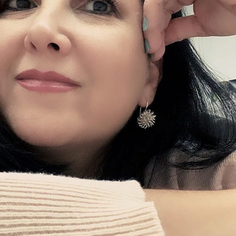 May include: A close-up of a person wearing a silver earring. The earring is a small, round design with a textured, floral-like pattern. The person has dark hair and is wearing a light-colored sweater.