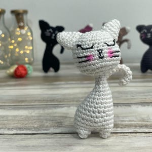 Può includere: Un gatto bianco all'uncinetto con occhi neri e guance rosa. Il gatto è in piedi su una superficie di legno.