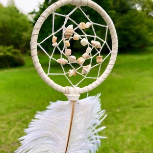 May include: Cream-colored dreamcatcher with a circular frame wrapped in fabric. The web is strung with white thread and adorned with small seashells and beads. A large white feather hangs below, against a backdrop of green grass and trees.