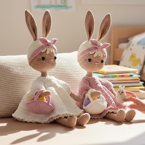 Könnte beinhalten: Zwei gehäkelte Hasenpuppen, eine in Weiß und die andere in Rosa, jeweils mit einem Korb. Die Puppen haben lange Ohren, Stirnbänder und Kleider. Sie sitzen auf einer hellen Oberfläche. Eine kleine Hasenfigur ist ebenfalls vorhanden.