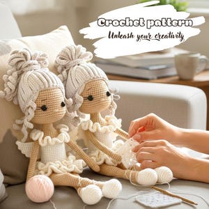 Può includere: Due bambole all'uncinetto fatte a mano con pelle marrone chiaro, abiti bianchi e capelli grigi ricci siedono su un divano grigio. Le bambole hanno occhi neri e ali bianche. L'immagine include il testo "Crochet pattern. Unleash your creativity."