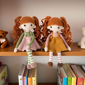 May include: Two handmade crochet dolls with red hair and dresses, one pink and one mustard yellow, sit on a wooden shelf. A brown teddy bear and a white teddy bear are also on the shelf. Books are visible below the shelf.