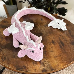 May include: A pink and white crocheted dragon toy with a long tail. The dragon is lying on a wooden table.