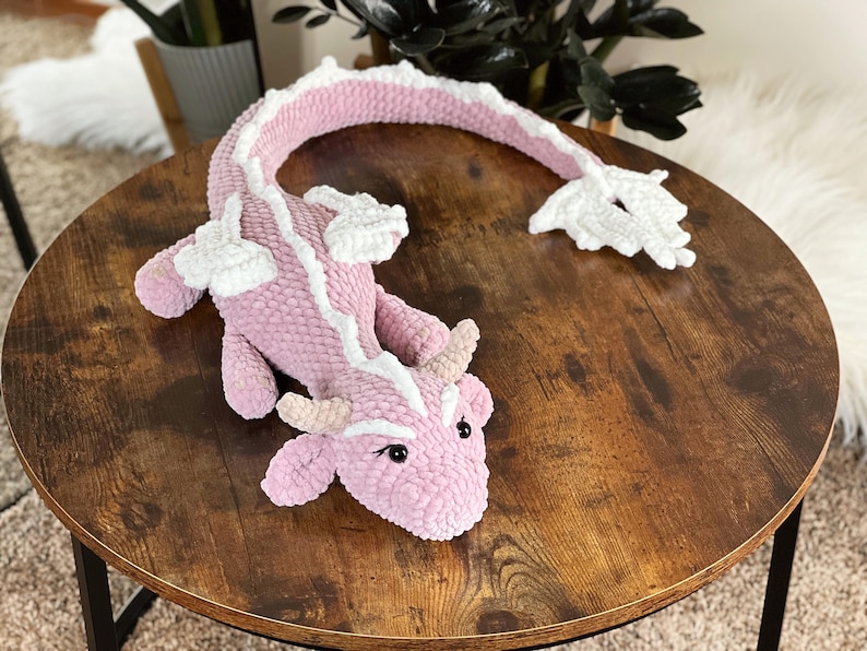 May include: A pink and white crocheted dragon toy with a long tail. The dragon is lying on a wooden table.