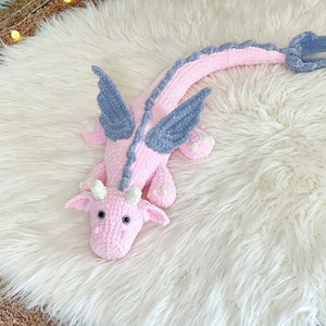 May include: A pink crocheted dragon with blue wings. The dragon is lying on a white fluffy rug.