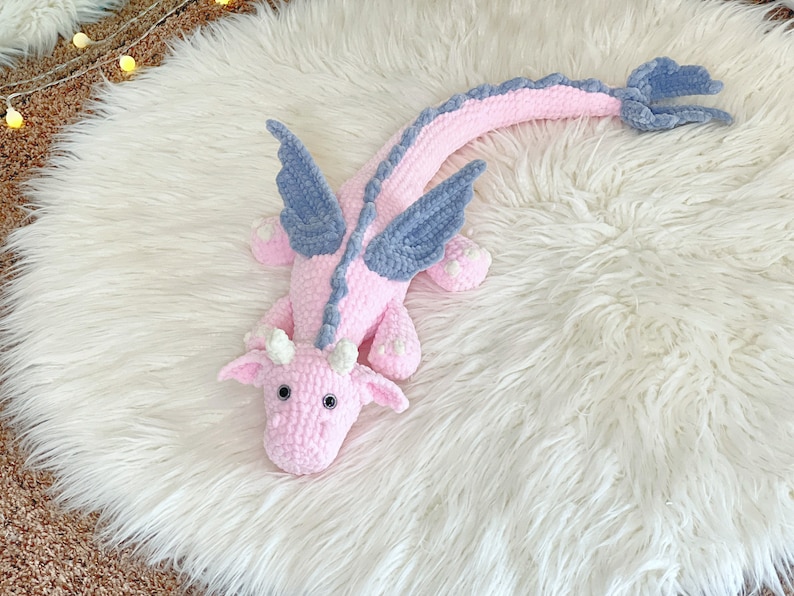 May include: A pink crocheted dragon with blue wings. The dragon is lying on a white fluffy rug.