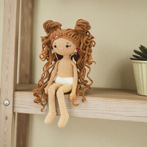 May include: A handmade crochet doll with light tan skin, wearing white underwear. The doll features long, curly brown hair styled in two top knots, accented with small colourful beads. The doll has embroidered facial features and sits upright.
