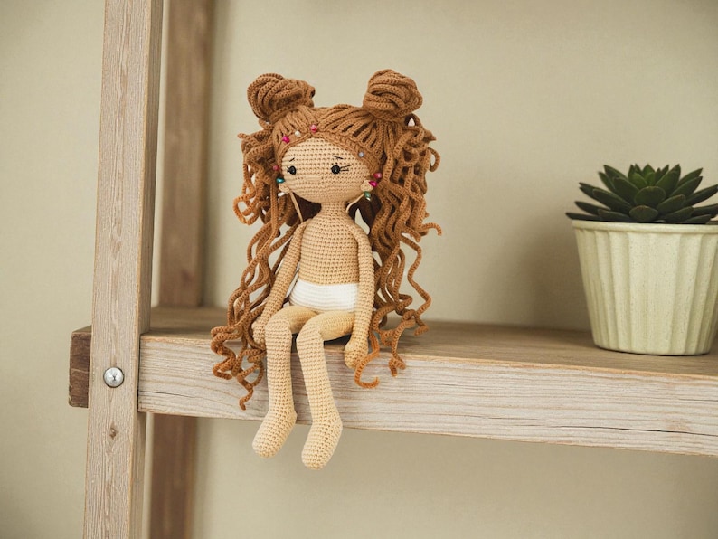 May include: A handmade crochet doll with light tan skin, wearing white underwear. The doll features long, curly brown hair styled in two top knots, accented with small colourful beads. The doll has embroidered facial features and sits upright.