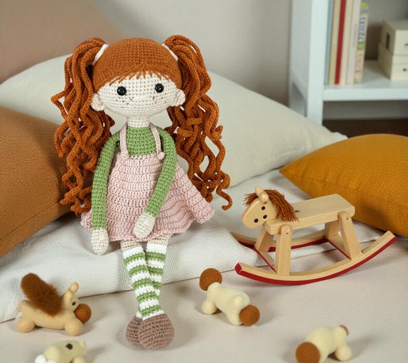 May include: A handmade crochet doll with red hair in pigtails, wearing a green top and pink dress. The doll has striped green and white legs and brown shoes. A wooden rocking horse and small toy animals are in the foreground.