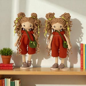 May include: Two identical handmade crochet dolls with long curly brown yarn hair styled in double buns with lime green accents. Each doll wears a rust-coloured jumpsuit and a small green crossbody bag, standing on circular wooden bases.