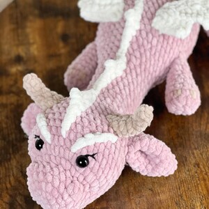 May include: A pink crocheted dragon with white accents. The dragon has a long tail, wings, and a small head with black eyes.