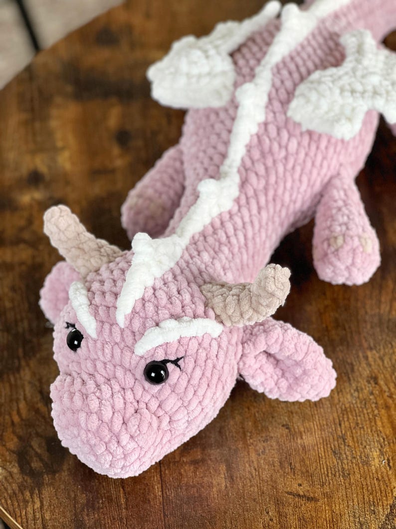 May include: A pink crocheted dragon with white accents. The dragon has a long tail, wings, and a small head with black eyes.