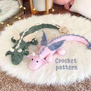 May include: Two crocheted dragon plushies, one green and one pink, with blue wings, on a white fluffy surface. The text "Crochet pattern" is written below the plushies.
