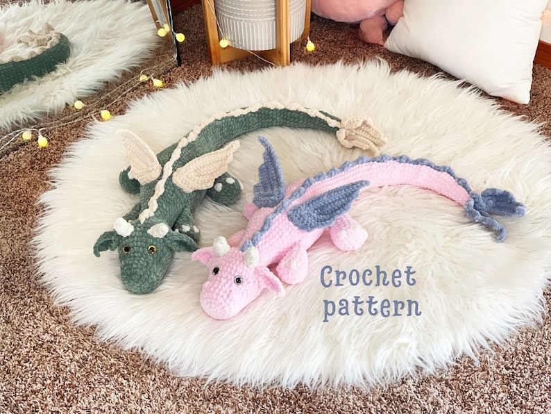 May include: Two crocheted dragon plushies, one green and one pink, with blue wings, on a white fluffy surface. The text "Crochet pattern" is written below the plushies.