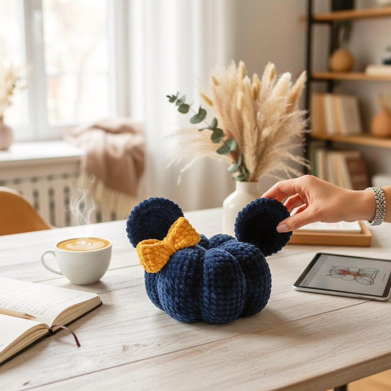 May include: A plush, dark blue pumpkin-shaped decor item featuring two round ears and a textured yellow bow attached to the front. The object is crafted from a soft, velvet-like material with visible stitching segments creating a rounded, segmented form.