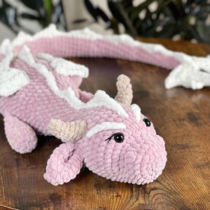 May include: A pink and white crocheted dragon plush toy with a long tail. The dragon has a friendly face with black eyes and a pink nose.