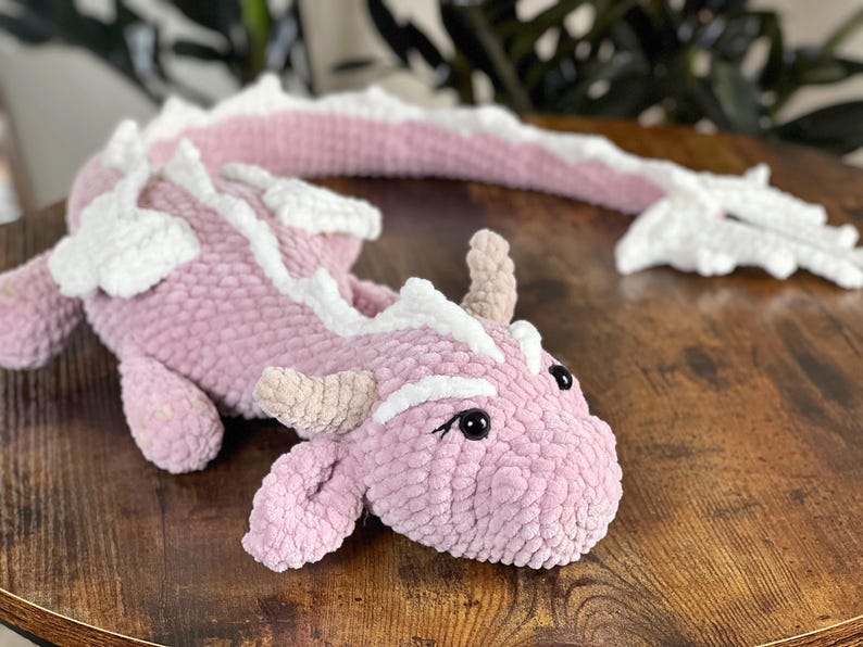 May include: A pink and white crocheted dragon plush toy with a long tail. The dragon has a friendly face with black eyes and a pink nose.
