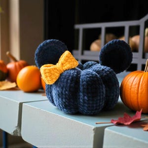 May include: A dark blue plush pumpkin with a textured, velvet-like finish, featuring two rounded ears and a bright yellow crocheted bow attached to the top. The pumpkin has a segmented, rounded shape and is positioned among several orange pumpkins.