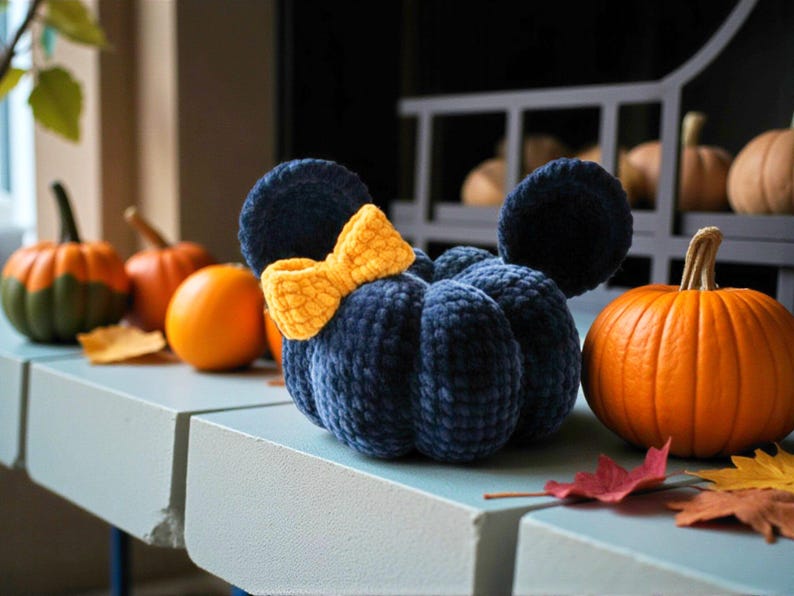 May include: A dark blue plush pumpkin with a textured, velvet-like finish, featuring two rounded ears and a bright yellow crocheted bow attached to the top. The pumpkin has a segmented, rounded shape and is positioned among several orange pumpkins.