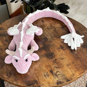 May include: A pink and white crocheted dragon plush toy with a curled tail. The dragon has a white snout and white accents on its back.