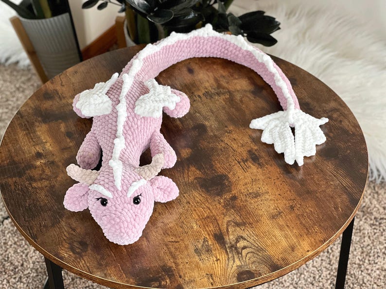 May include: A pink and white crocheted dragon plush toy with a curled tail. The dragon has a white snout and white accents on its back.