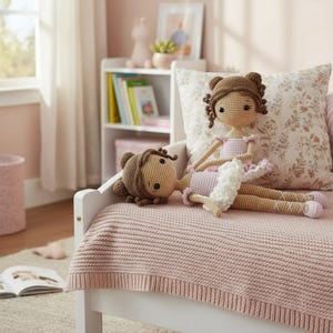 Puede incluir: Dos mu&ntilde;ecas de bailarina de ganchillo hechas a mano. Una mu&ntilde;eca est&aacute; de pie, la otra est&aacute; acostada. Ambas mu&ntilde;ecas tienen cabello casta&ntilde;o, tut&uacute;s rosas y zapatillas de ballet. Descansan sobre una manta rosa tejida en un marco de cama blanco.