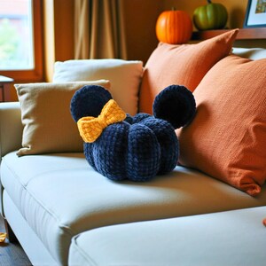 May include: A dark navy blue plush pumpkin-shaped cushion featuring two rounded ears and a bright yellow crocheted bow attached to the side. The cushion has a soft, textured surface with visible stitching segments that define its rounded shape.