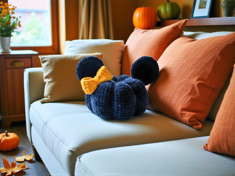 May include: A dark navy blue plush pumpkin-shaped cushion featuring two rounded ears and a bright yellow crocheted bow attached to the side. The cushion has a soft, textured surface with visible stitching segments that define its rounded shape.