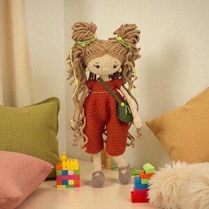 May include: A handmade crochet doll with long, curly light brown hair styled in two high buns with light green ribbons. The doll wears a rust-coloured short-sleeved jumpsuit, a small dark green crossbody bag, and grey shoes, featuring embroidered facial features.