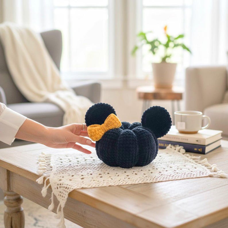 May include: A dark navy blue crocheted pumpkin-shaped plush toy featuring two round ears on top and a textured mustard yellow crocheted bow attached to the front. The object is crafted with a soft, ribbed yarn texture and a rounded, segmented form.
