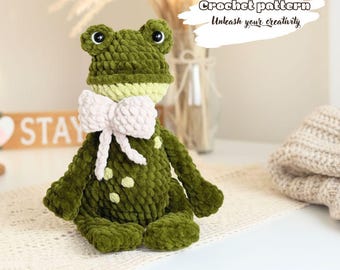 May include: A handmade green crochet frog with a light pink bow tie. The frog has white polka dots and large black eyes. The text "Crochet pattern Unleash your creativity" is visible in the background.