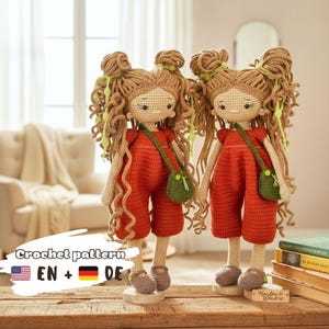 May include: Two handmade crochet dolls with long curly brown yarn hair styled in double buns, wearing orange jumpsuits and small green crossbody bags. The dolls have light beige skin, embroidered facial features, and small brown shoes.