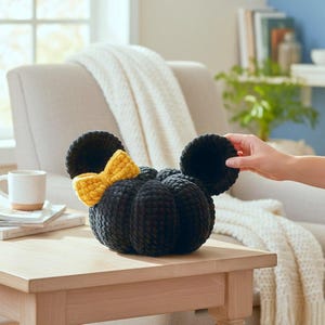 May include: A black crocheted pumpkin-shaped plush with two large round black ears and a bright yellow crocheted bow attached to the side. The plush features a soft, textured yarn construction with a segmented, rounded body and a plush bow.