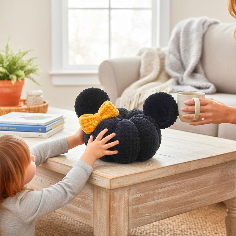 May include: A black crocheted pumpkin-shaped plush toy featuring two round ears and a bright yellow crocheted bow attached to the front. The plush has a textured, yarn-based surface and is positioned on a wooden table surface.