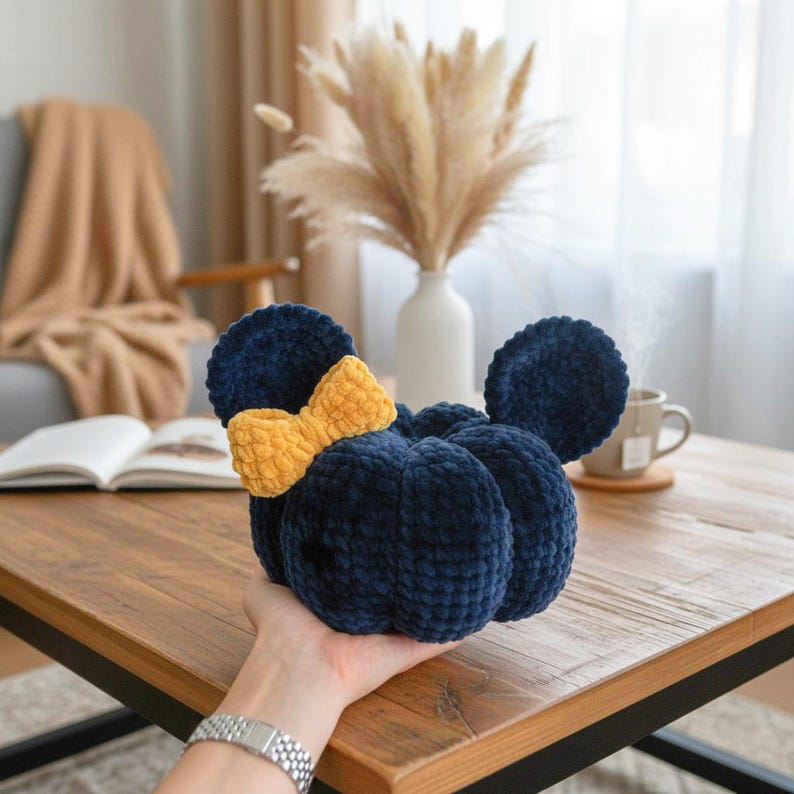 May include: A plush, dark blue pumpkin-shaped decorative item featuring two rounded ears and a textured yellow bow attached to the side. The object is crafted from a soft, velvety material with a segmented, rounded design, resembling a character silhouette.