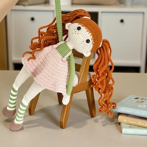 May include: A handmade crochet doll with red hair, wearing a pink dress and green accents, is suspended in the air. The doll is seated on a small wooden chair. The doll's legs have green and white striped socks and brown shoes.
