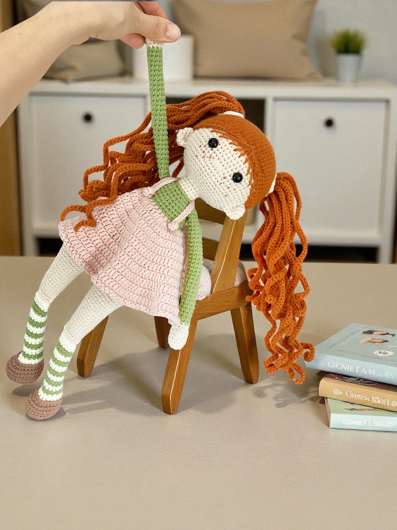 May include: A handmade crochet doll with red hair, wearing a pink dress and green accents, is suspended in the air. The doll is seated on a small wooden chair. The doll's legs have green and white striped socks and brown shoes.