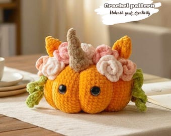 May include: A crocheted orange pumpkin plush with two black eyes, a central tan horn, and a crown of light pink and white crocheted flowers. Green crocheted vines extend from the sides. Text above reads: Crochet pattern Unleash your creativity.