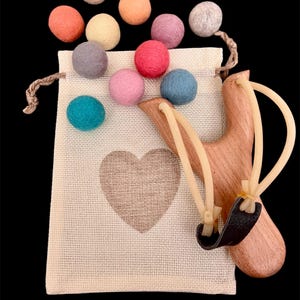 K&ouml;nnte beinhalten: Eine Holzschleuder mit schwarzem Lederbeutel und Gummib&auml;ndern, neben einem beigen Kordelzugbeutel mit Herzmotiv. Bunte Filzkugeln in Rosa, Blau, Gr&uuml;n, Orange und Grau sind um die Tasche herum verstreut.