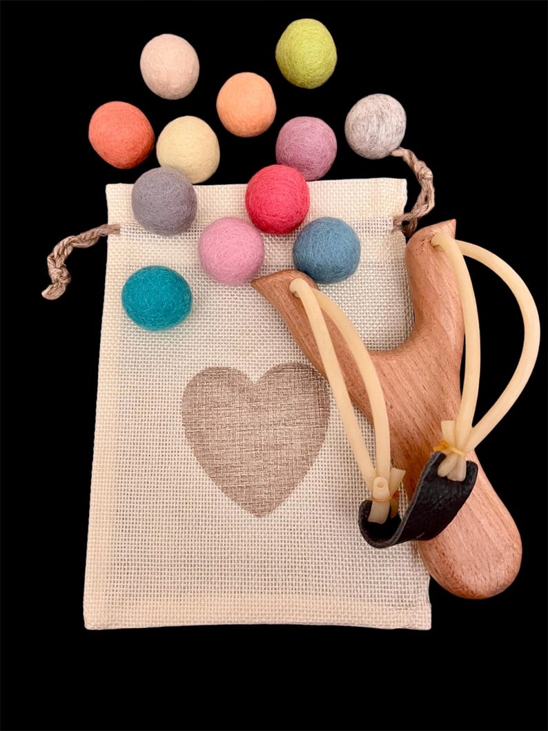 K&ouml;nnte beinhalten: Eine Holzschleuder mit schwarzem Lederbeutel und Gummib&auml;ndern, neben einem beigen Kordelzugbeutel mit Herzmotiv. Bunte Filzkugeln in Rosa, Blau, Gr&uuml;n, Orange und Grau sind um die Tasche herum verstreut.