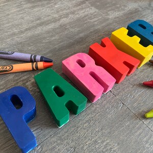 May include: The name "Parker" spelled out in colorful crayon letters. The letters are arranged in a line from left to right. The letters are made of different colors, including blue, green, pink, red, yellow, and blue. There are also several crayons lying on the wooden surface.
