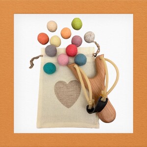 K&ouml;nnte beinhalten: Eine Holzschleuder mit einer schwarzen Ledertasche und Gummib&auml;ndern, neben einem beigefarbenen Stoffbeutel mit einem Herzmotiv. Der Beutel ist mit bunten Filzkugeln in Orange-, Gr&uuml;n-, Rosa- und Blaut&ouml;nen gef&uuml;llt.
