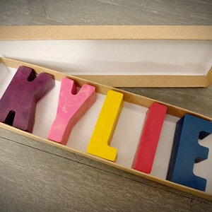 May include: A cardboard box with a white interior, containing colorful wax letters spelling out the name "KYLIE". The letters are in shades of purple, pink, yellow, red, and blue.