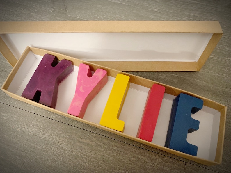 May include: A cardboard box with a white interior, containing colorful wax letters spelling out the name "KYLIE". The letters are in shades of purple, pink, yellow, red, and blue.