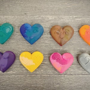 May include: Eight heart-shaped crayons with different colors and facial expressions. The crayons are arranged in two rows of four. The colors are teal, blue, brown, orange, purple, yellow, pink, and gray.