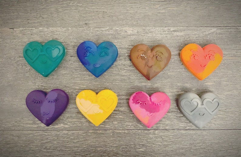 May include: Eight heart-shaped crayons with different colors and facial expressions. The crayons are arranged in two rows of four. The colors are teal, blue, brown, orange, purple, yellow, pink, and gray.