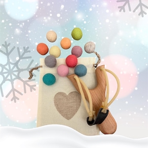 K&ouml;nnte beinhalten: Eine Holzschleuder mit einem schwarzen Lederbeutel und beigen Gummib&auml;ndern. Ein beiger Stoffbeutel mit Herzmuster ist mit bunten Filzkugeln gef&uuml;llt. Der Hintergrund hat ein Winterthema mit Schneeflocken und Bokeh.
